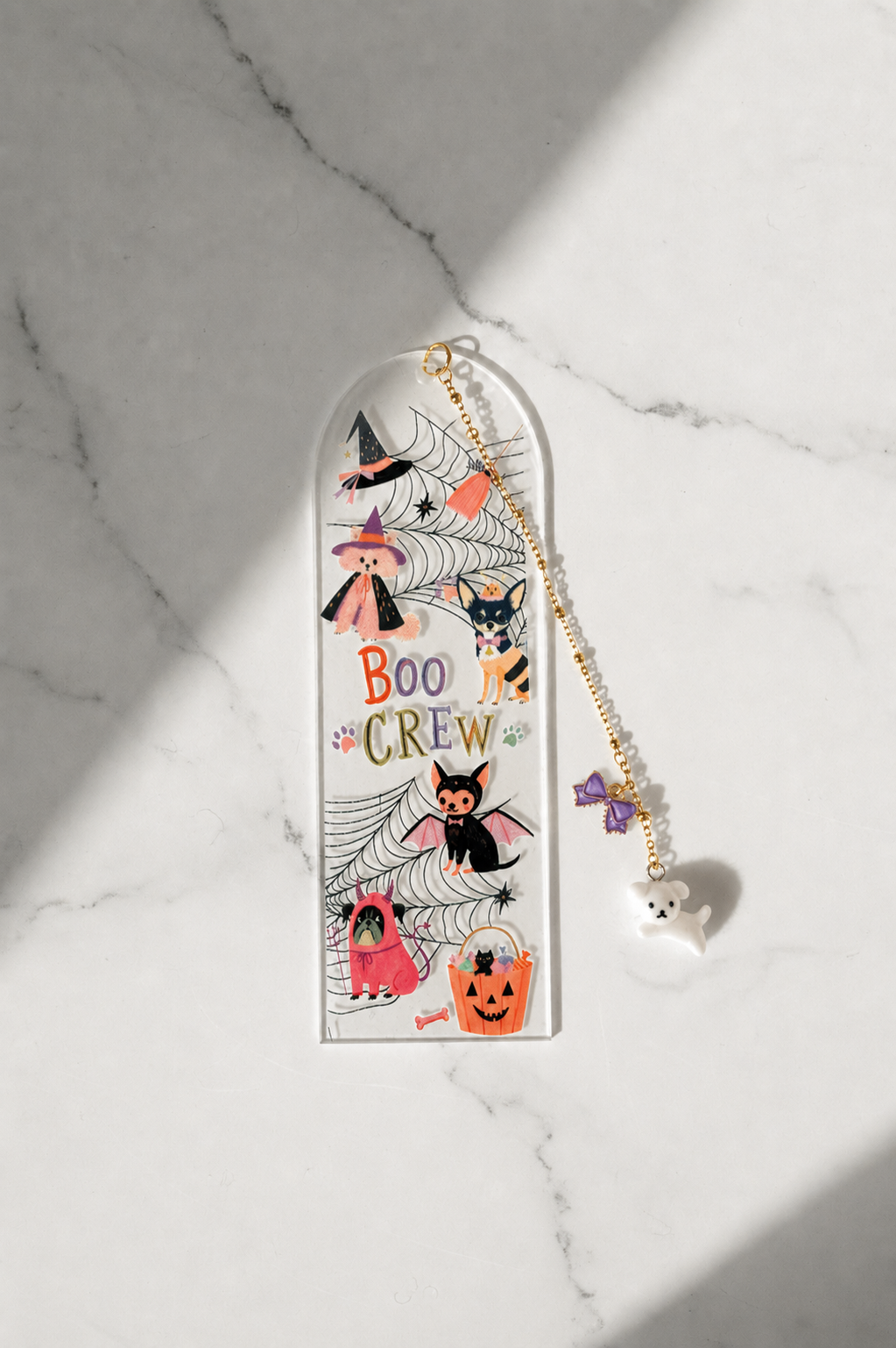 A Halloween-themed acrylic bookmark with a gold-tone chain and ghost charm, displayed on a white marble surface with soft diagonal shadows.