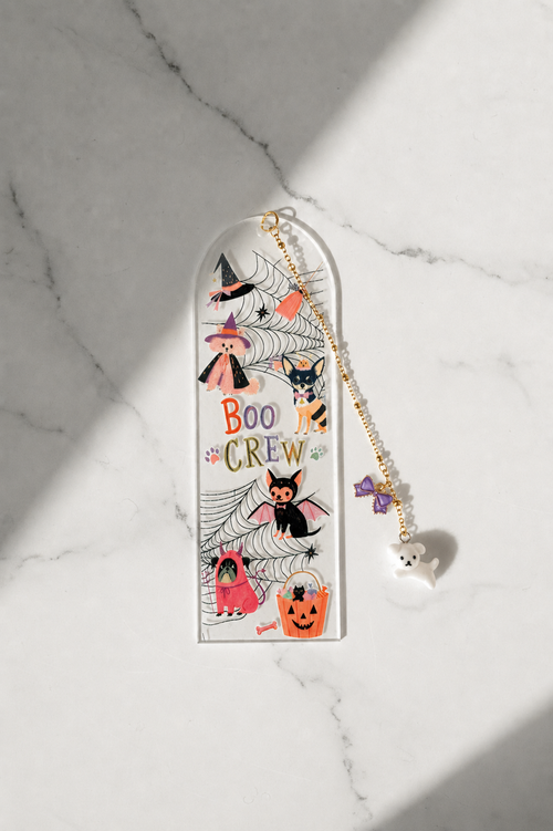 A Halloween-themed acrylic bookmark with a gold-tone chain and ghost charm, displayed on a white marble surface with soft diagonal shadows.