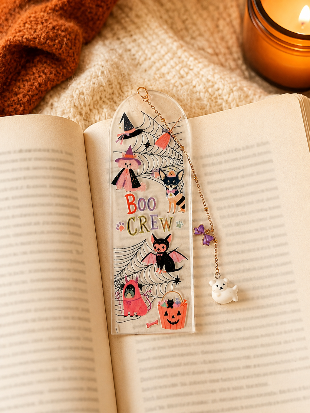 A Halloween-themed clear acrylic bookmark with a gold chain and charms, resting on an open book. The bookmark features illustrations of a witch, cat, spider, ghost, and pumpkin with the text 'BOO CREW'.