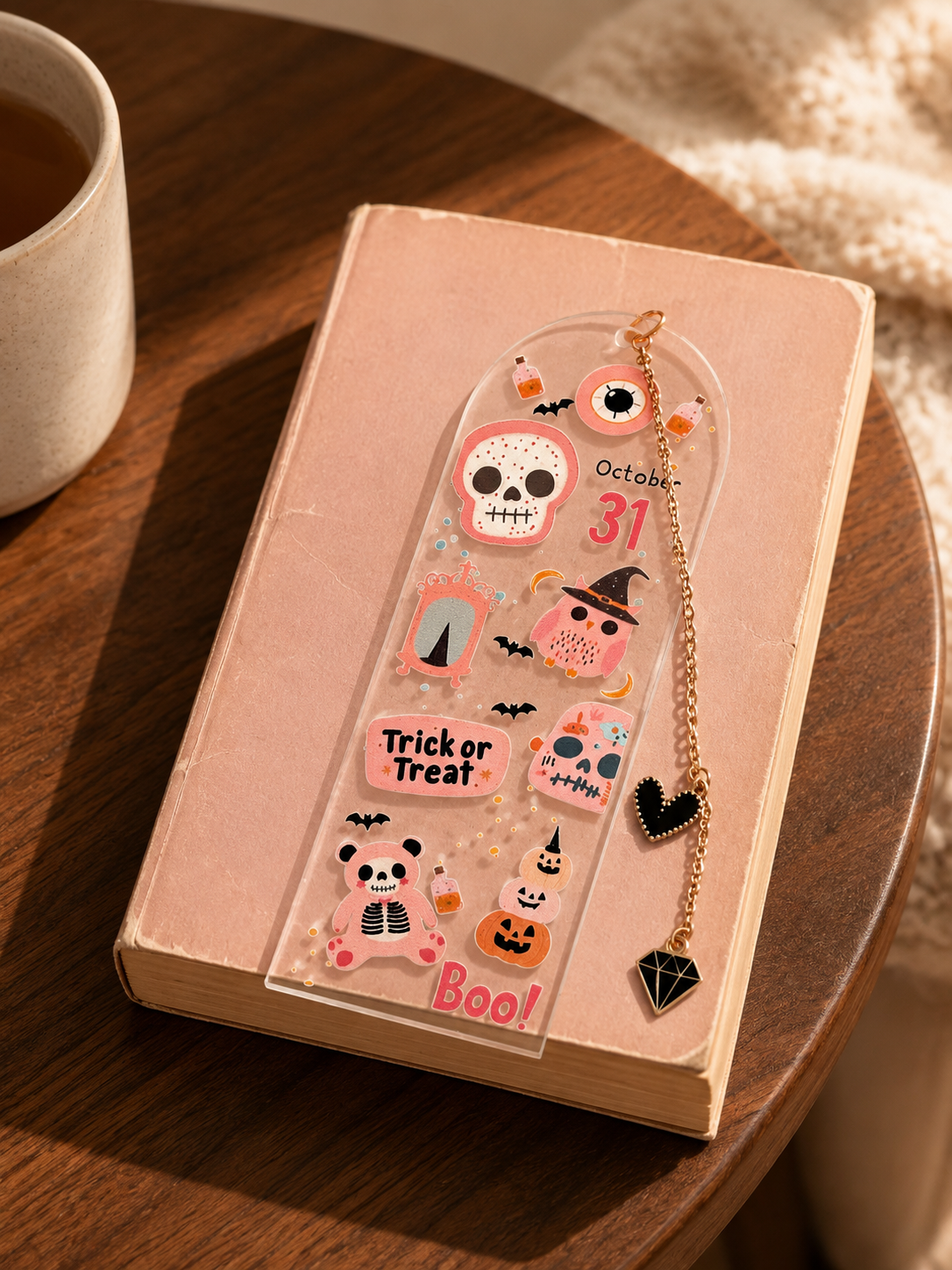 A clear acrylic Halloween-themed bookmark or page divider with pink and black enamel details, resting on a pink book on a wooden table next to a mug.