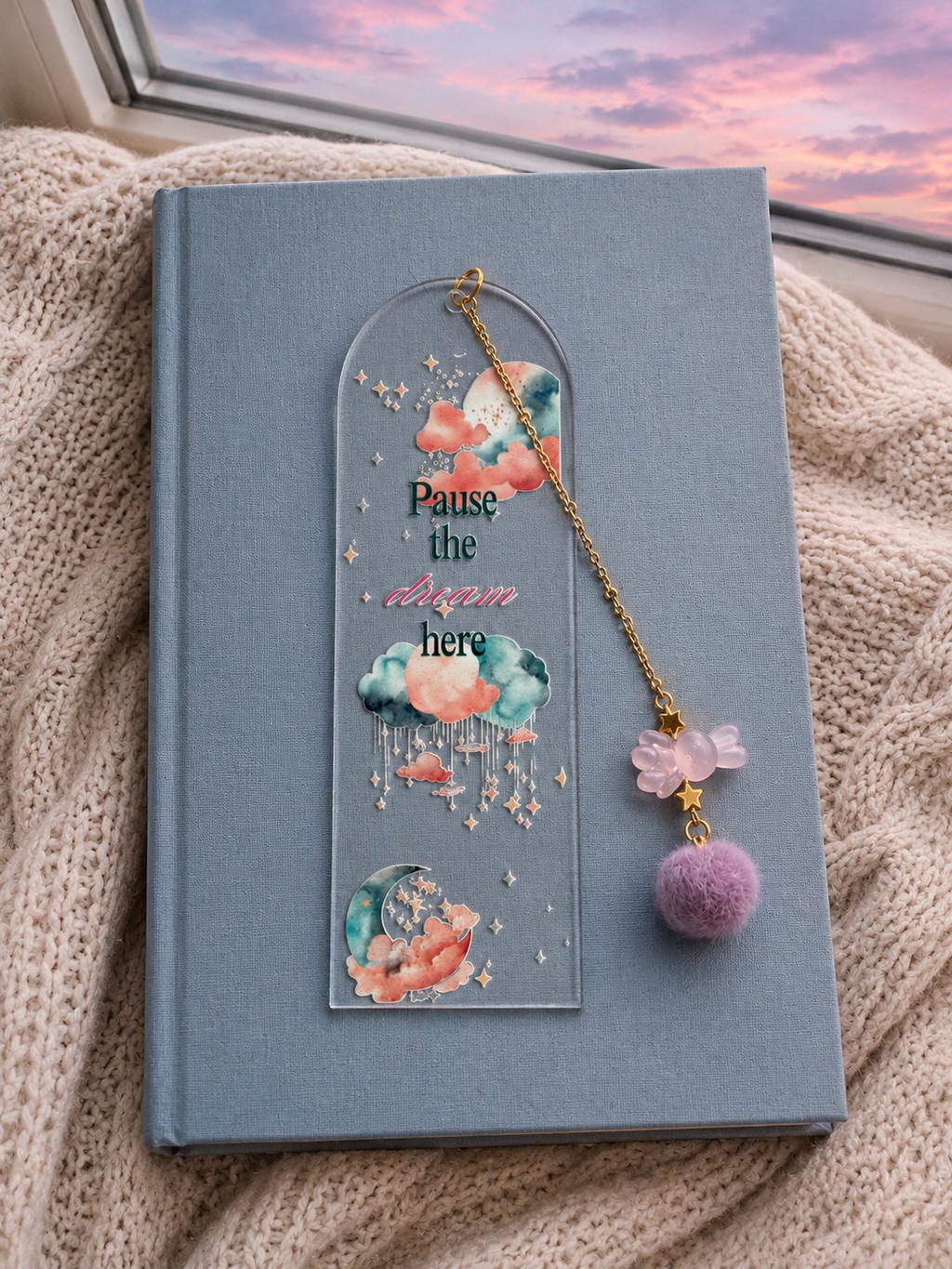 A decorative acrylic bookmark with a gold chain and purple pom-pom charm rests on a blue hardcover book, placed on a beige knit blanket near a window with a pink sunset sky visible.