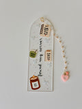 A clear acrylic bookmark with a gold loop at the top, decorated with cute illustrations of candles, a green cup, notebook, and the phrase 'found my corner' in black script. It features a dangling beaded chain with a small pink heart charm on a light gray background.