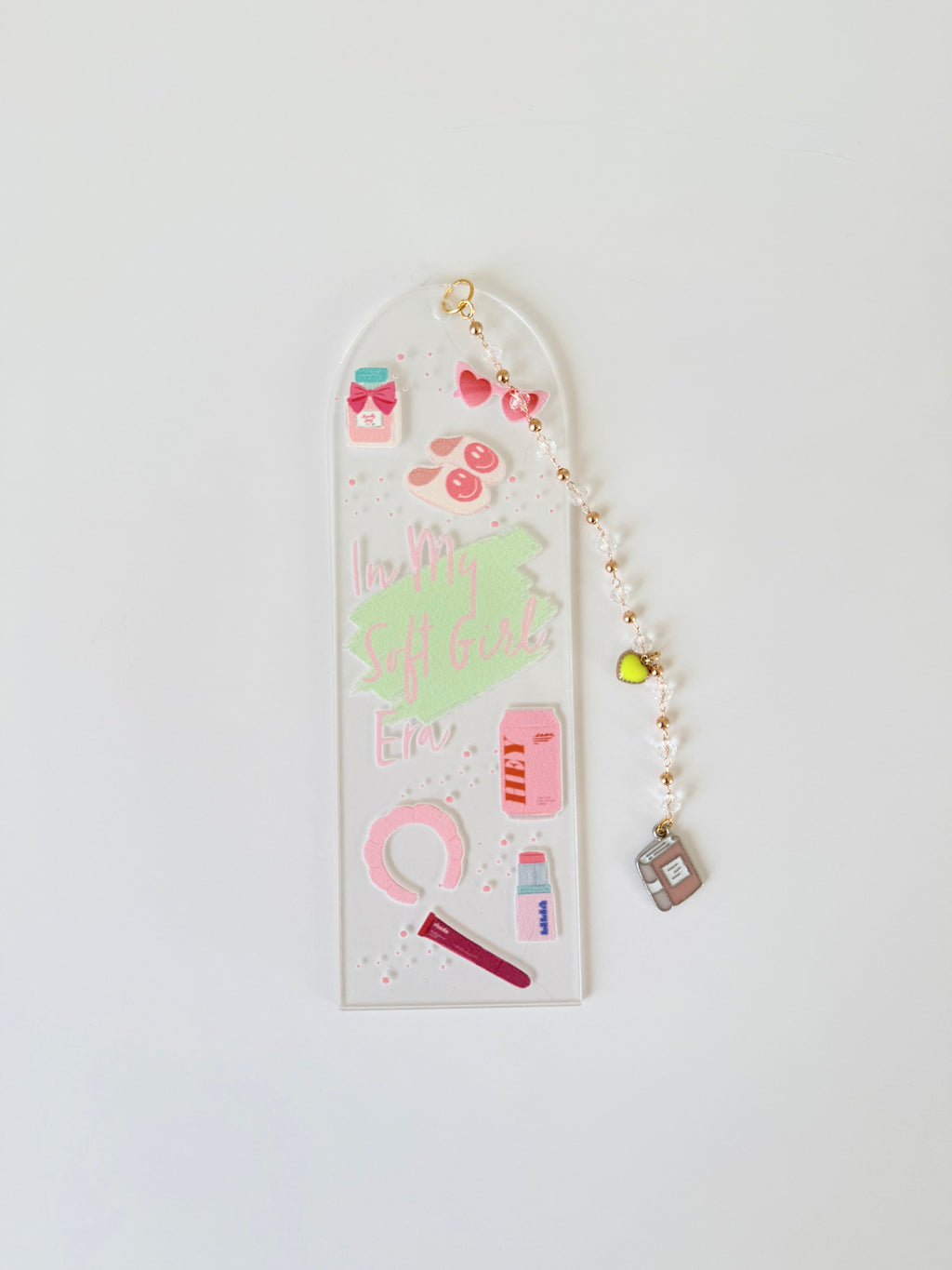 A clear acrylic bookmark with a rounded top, decorated with cute pink and green stickers, gold glitter, and a dangling beaded charm with a small house, shown on a plain white background.