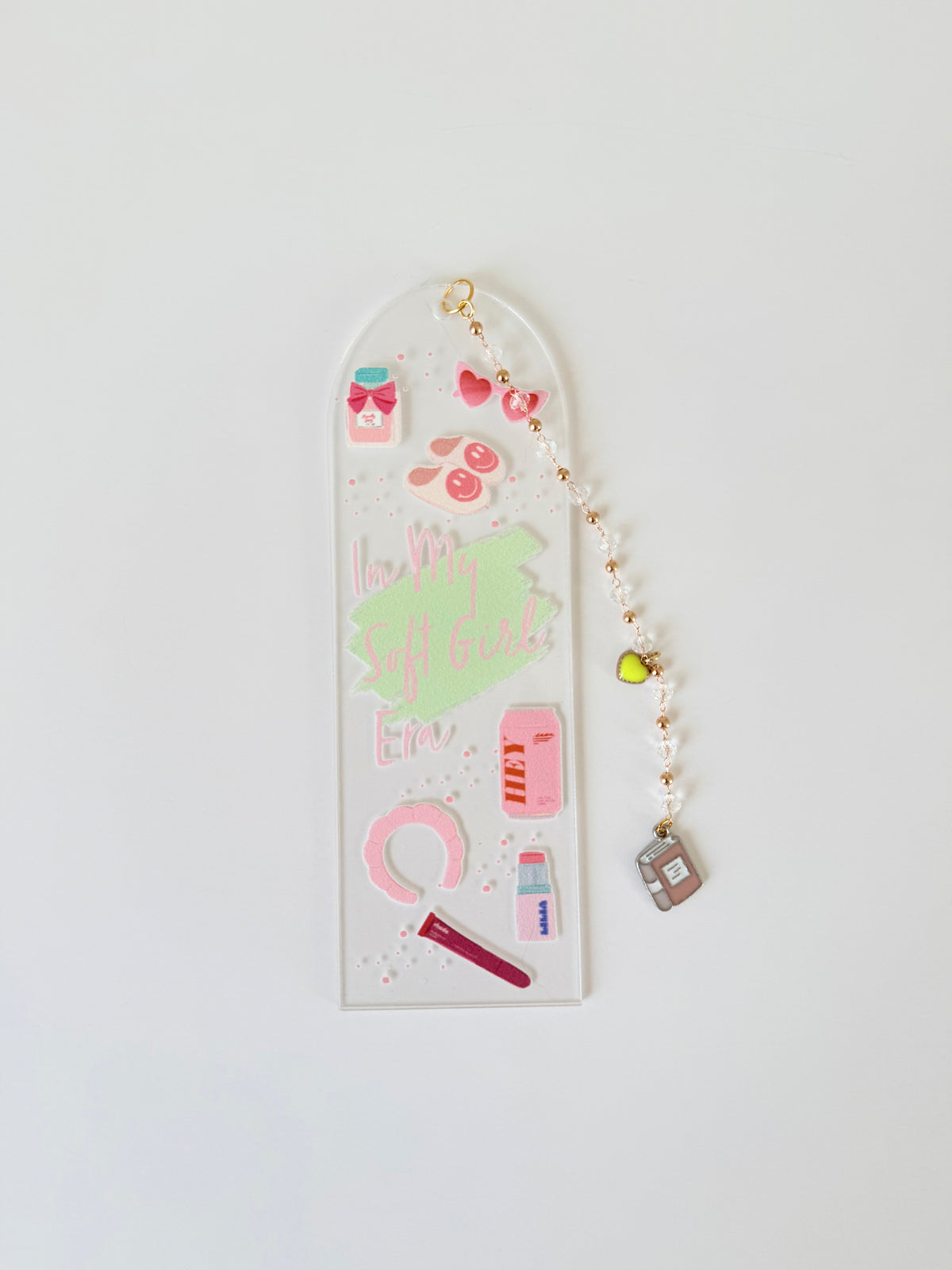 A clear acrylic bookmark with a rounded top, decorated with cute pink and green stickers, gold glitter, and a dangling beaded charm with a small house, shown on a plain white background.
