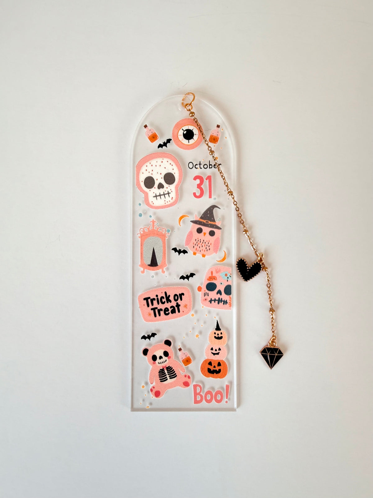 A clear acrylic Halloween bookmark with a rose gold chain and black heart pendant, decorated with cute cartoon illustrations including a skull, witch hat, jack-o'-lantern, and the words 'October 31', 'Trick or Treat', and 'Boo!'.