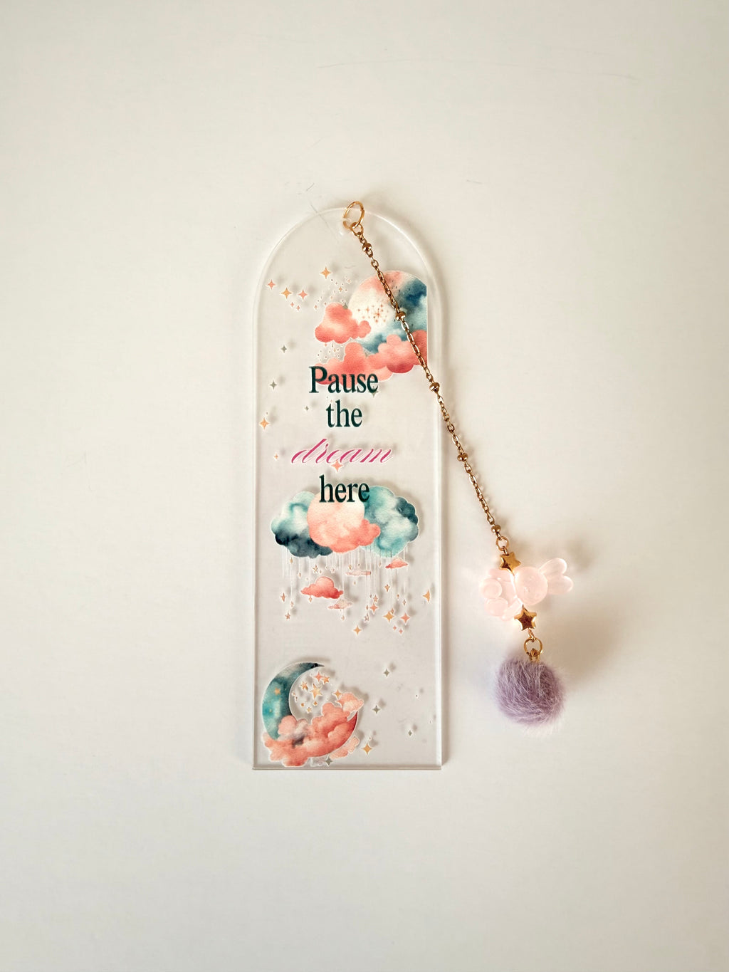 An arched clear acrylic wall hanging with watercolor clouds, gold glitter, and the text 'Pause the dream here,' shown with a dangling gold chain, pink flower charm, and lavender pom-pom against a plain light background.