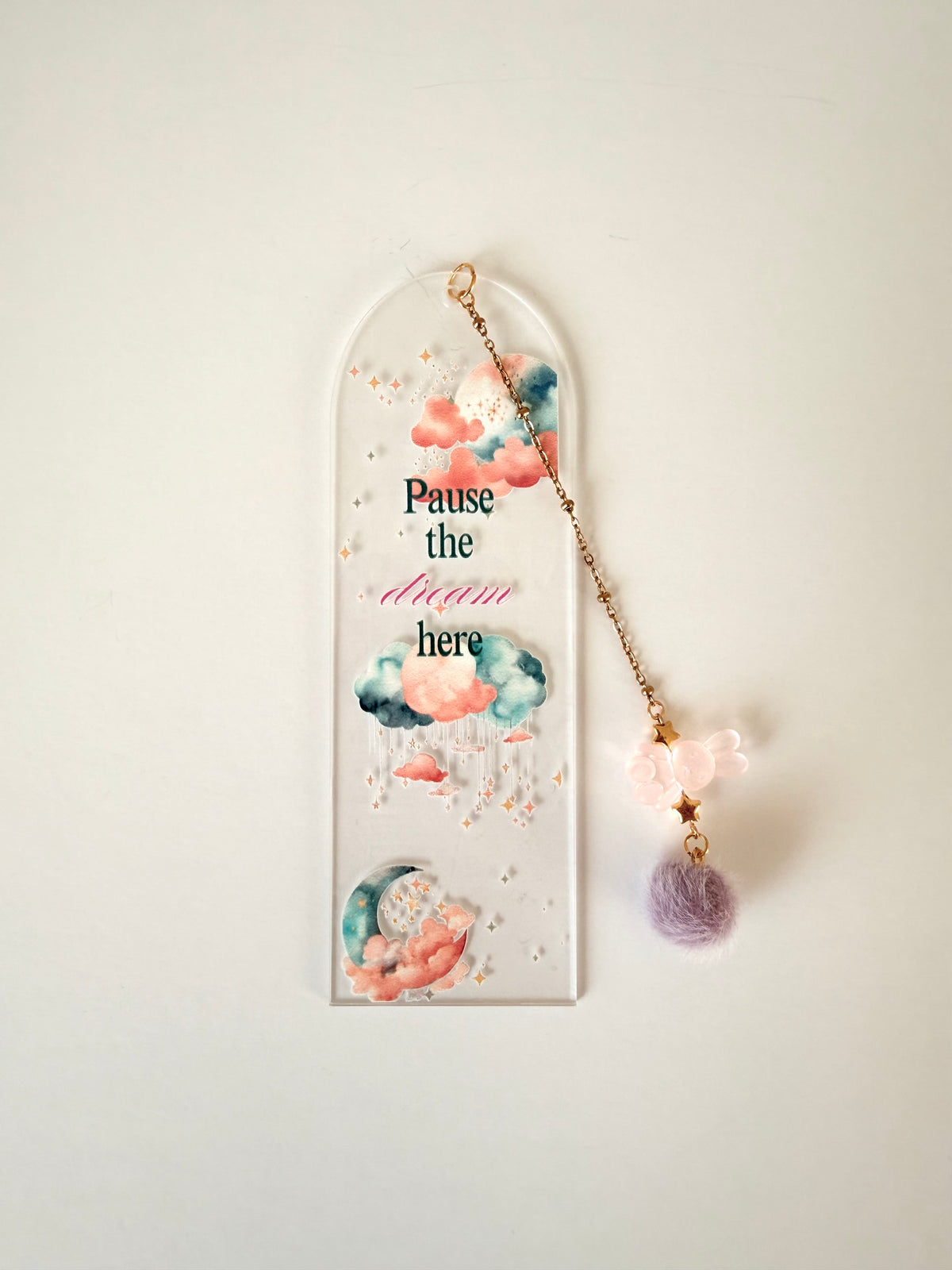 An arched clear acrylic wall hanging with watercolor clouds, gold glitter, and the text 'Pause the dream here,' shown with a dangling gold chain, pink flower charm, and lavender pom-pom against a plain light background.