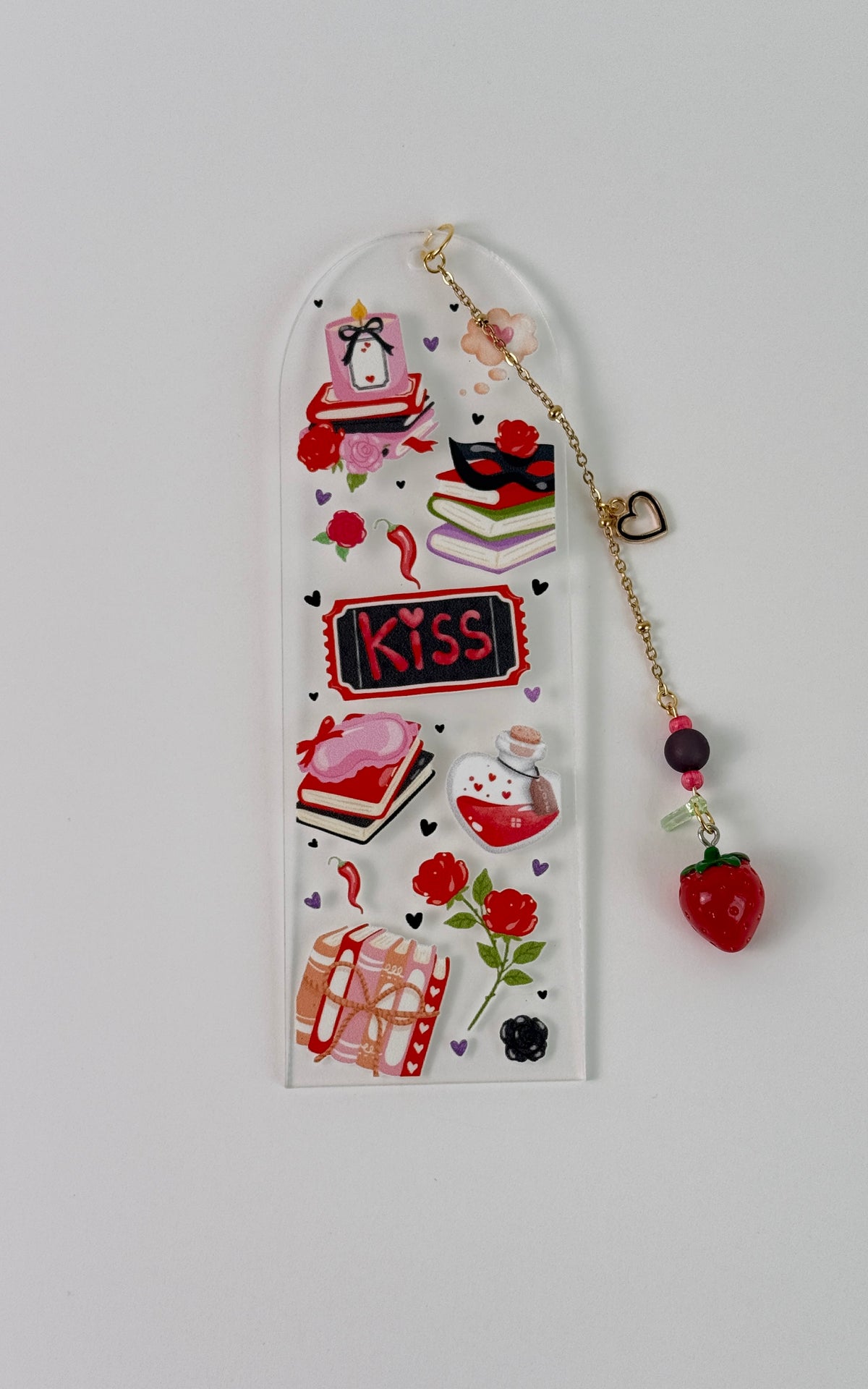 A arched acrylic bookmark with a red strawberry charm with gold colored chain. Has artwork that has candles, books, potion and roses. Laying on a white back ground. 