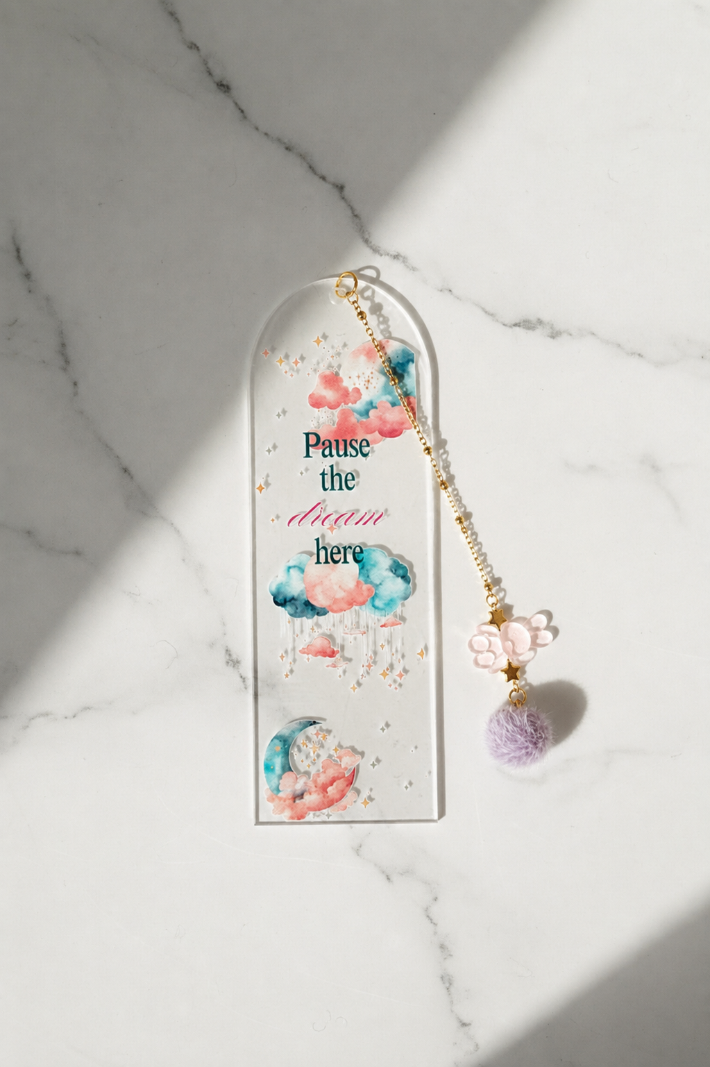 A clear acrylic arch-shaped bookmark with watercolor clouds, gold foil specks, and the text 'Pause the dream here,' hanging from a gold beaded chain with pink flower charms and a purple pom-pom, displayed on a white marble surface with diagonal shadows.