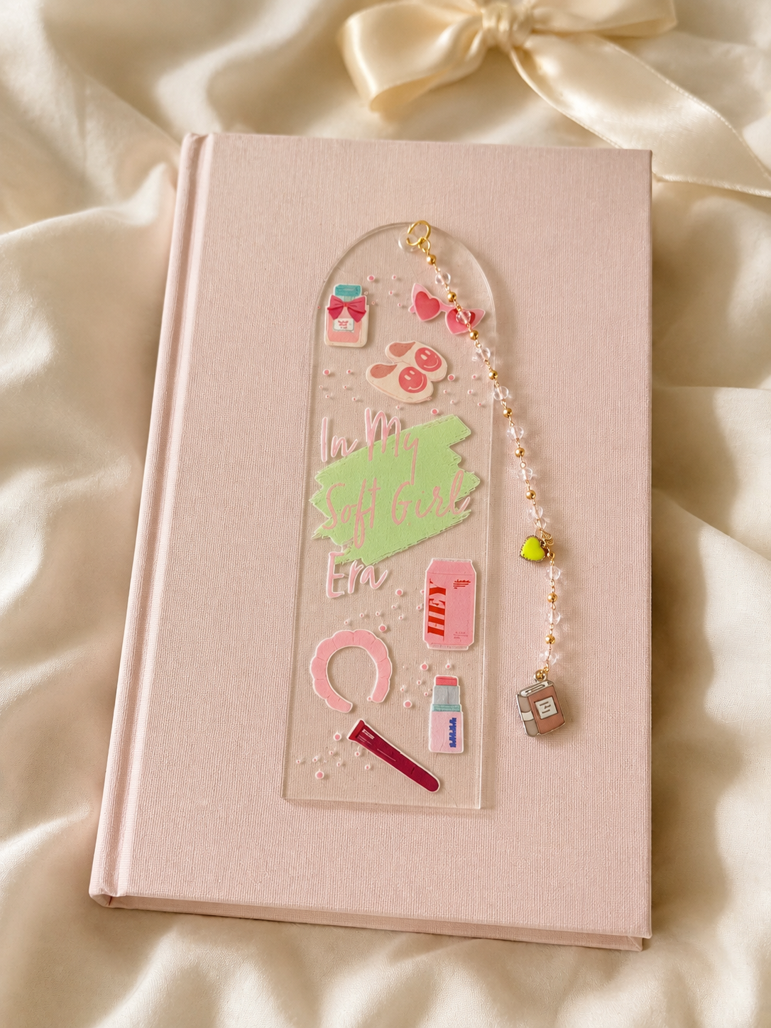 A clear acrylic phone strap with pink and green cosmetic-themed charms, displayed on a light pink hardcover notebook over draped cream satin fabric.