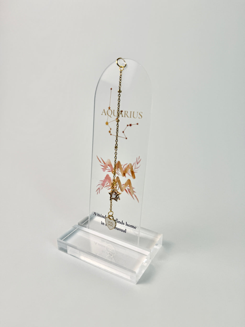 Clear arched acrylic bookmark with Aquarius zodiac lettering, constellation artwork, a pink-and-gold water-bearer symbol, the quote “A mind that finds home in the unusual,” and dangling star and Aquarius heart charms.