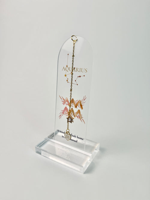 Clear arched acrylic bookmark with Aquarius zodiac lettering, constellation artwork, a pink-and-gold water-bearer symbol, the quote “A mind that finds home in the unusual,” and dangling star and Aquarius heart charms.