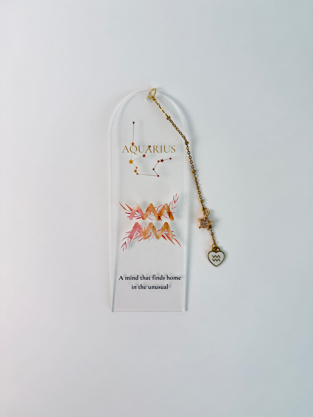 Clear arched acrylic bookmark with Aquarius zodiac lettering, constellation artwork, a pink-and-gold water-bearer symbol, the quote “A mind that finds home in the unusual,” and dangling star and Aquarius heart charms. on white background