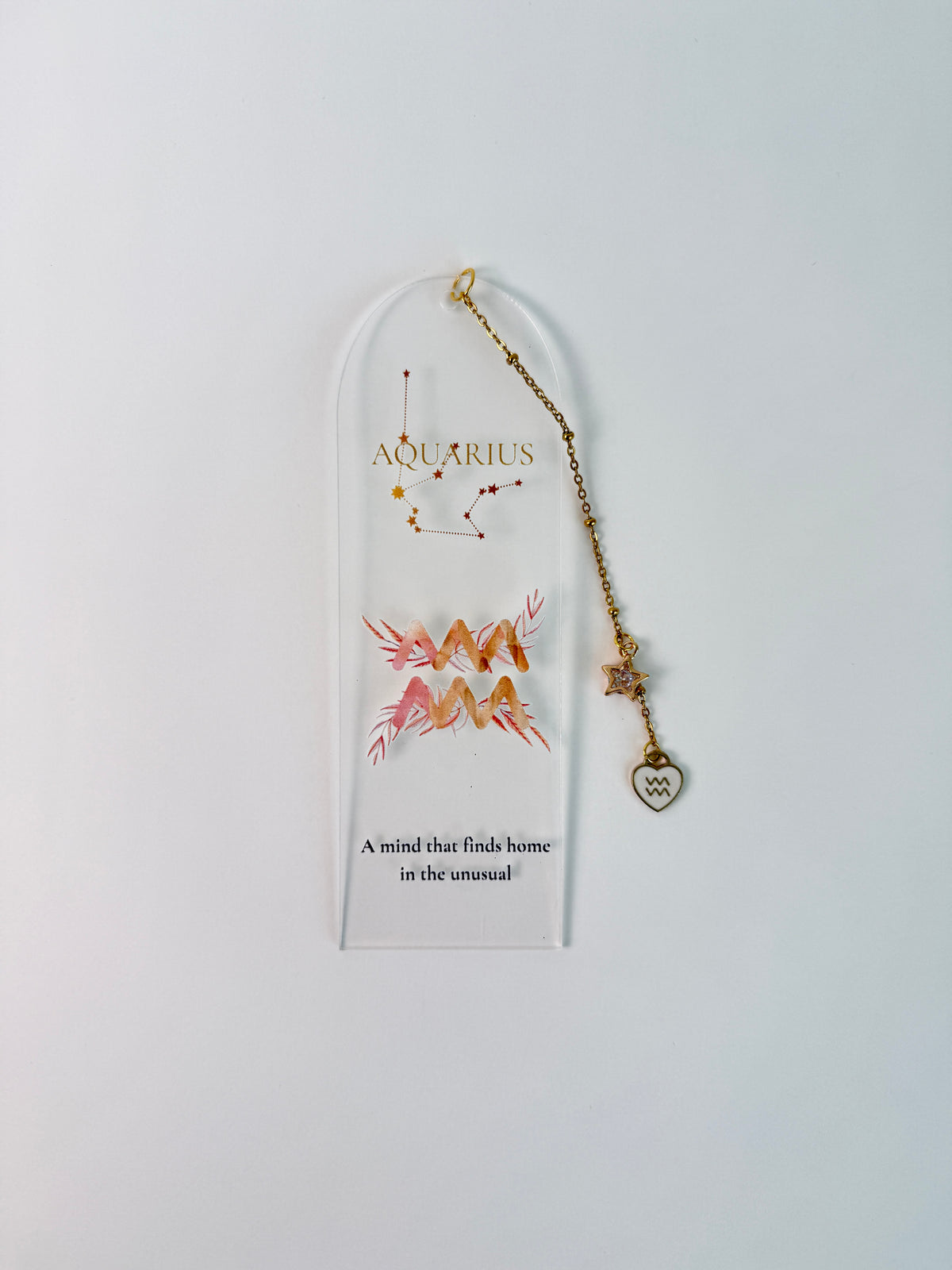Clear arched acrylic bookmark with Aquarius zodiac lettering, constellation artwork, a pink-and-gold water-bearer symbol, the quote “A mind that finds home in the unusual,” and dangling star and Aquarius heart charms. on white background