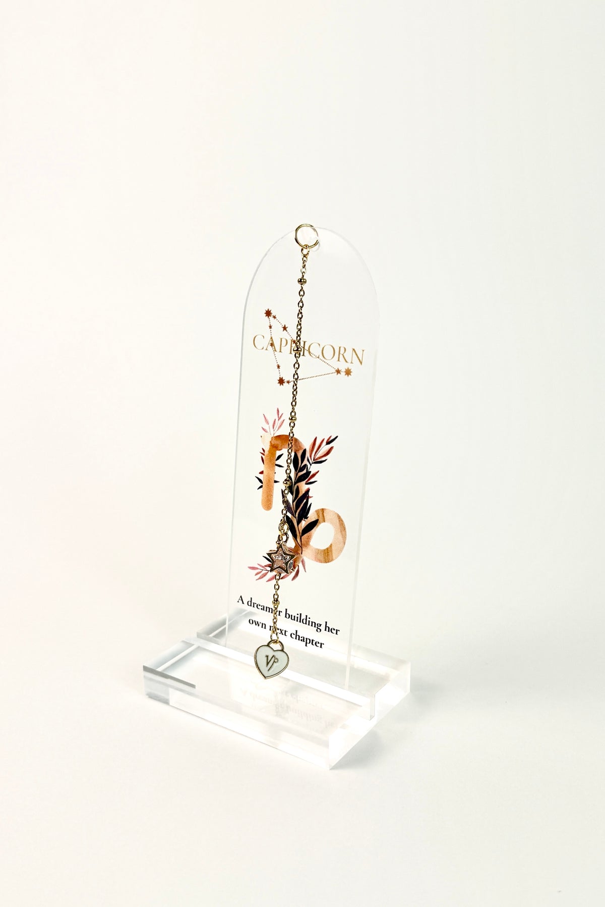 Clear arched acrylic bookmark with Capricorn zodiac lettering, constellation artwork, botanical details, the quote “A dreamer building her own next chapter,” and dangling star and Capricorn heart charms. standing on white background