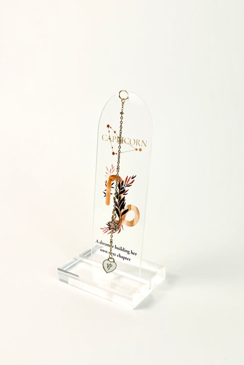 Clear arched acrylic bookmark with Capricorn zodiac lettering, constellation artwork, botanical details, the quote “A dreamer building her own next chapter,” and dangling star and Capricorn heart charms. standing on white background