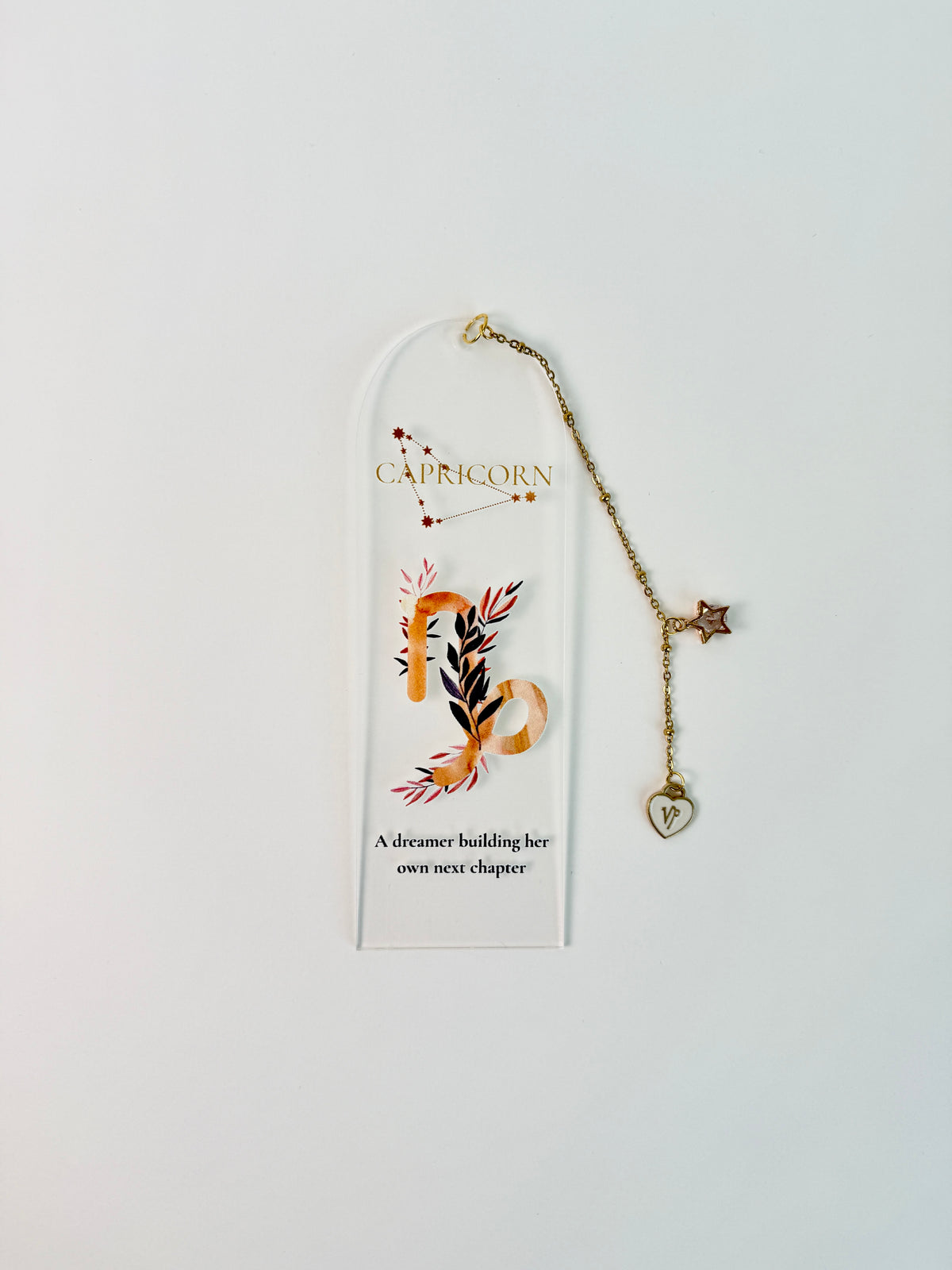 Clear arched acrylic bookmark with Capricorn zodiac lettering, constellation artwork, botanical details, the quote “A dreamer building her own next chapter,” and dangling star and Capricorn heart charms. laying flat on white background
