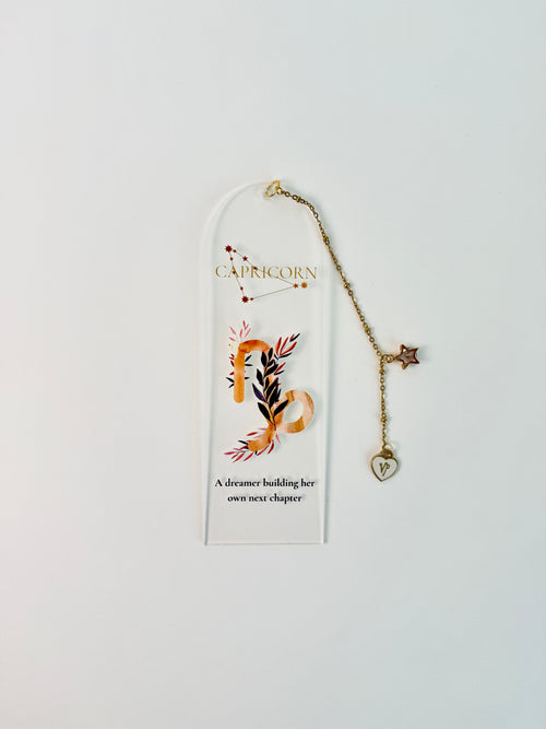 Clear arched acrylic bookmark with Capricorn zodiac lettering, constellation artwork, botanical details, the quote “A dreamer building her own next chapter,” and dangling star and Capricorn heart charms. laying flat on white background