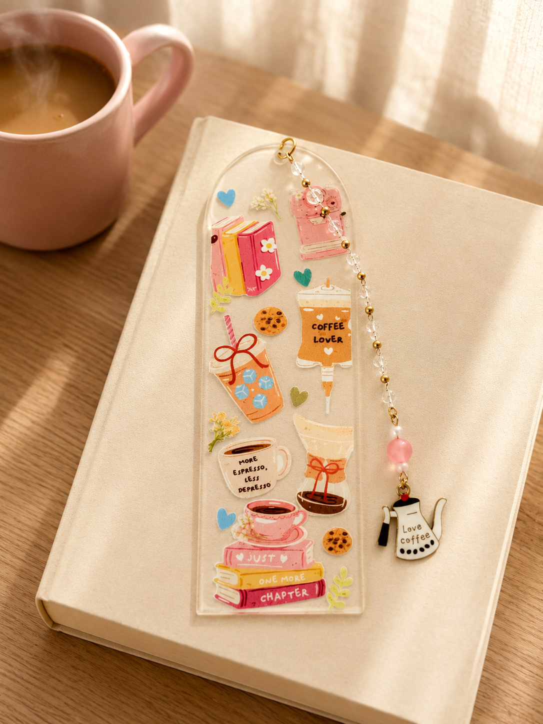 A clear acrylic bookmark with colorful coffee-themed illustrations and a dangling charm, resting on a cream-colored book on a wooden table next to a pink mug.