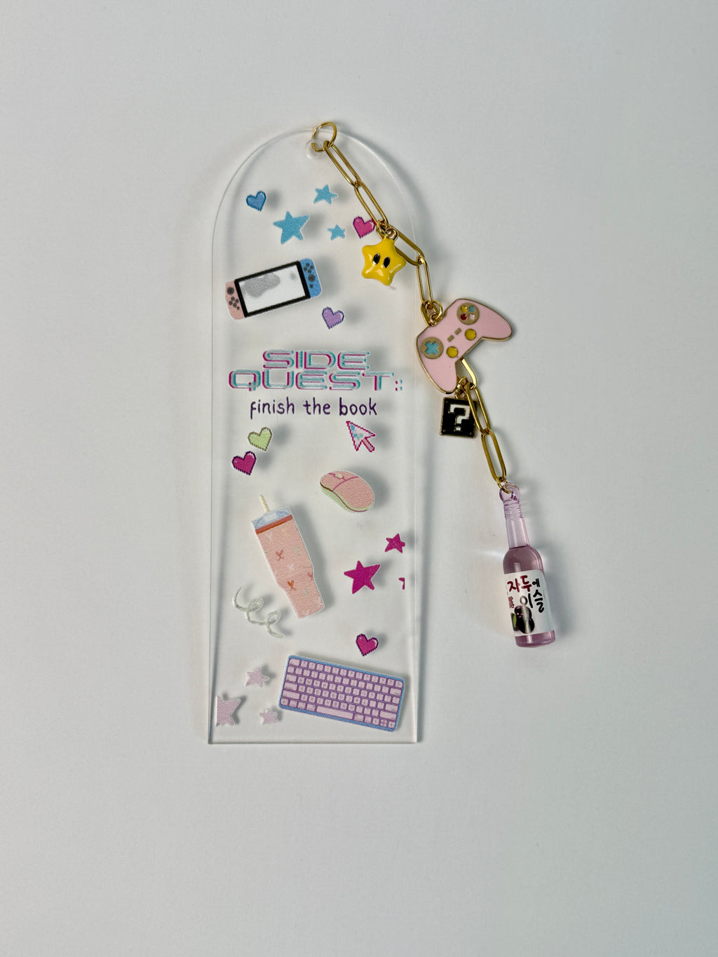 A clear acrylic keychain holder with the text 'SIDE QUEST: finish the book' and decorated with various colorful miniature gaming and tech-themed charms, including a keyboard, mouse, smartphone, controller, and a small bottle charm, shown against a plain light gray background.