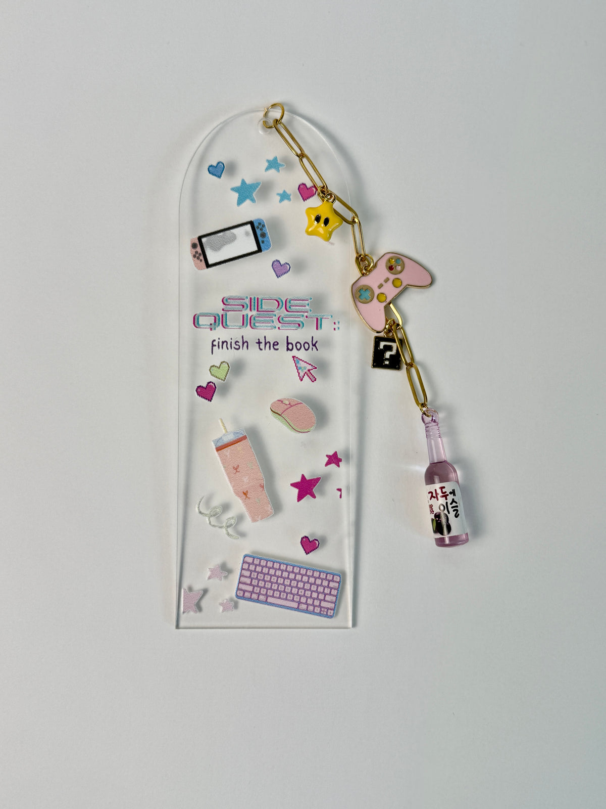 A clear acrylic keychain holder with the text 'SIDE QUEST: finish the book' and decorated with various colorful miniature gaming and tech-themed charms, including a keyboard, mouse, smartphone, controller, and a small bottle charm, shown against a plain light gray background.