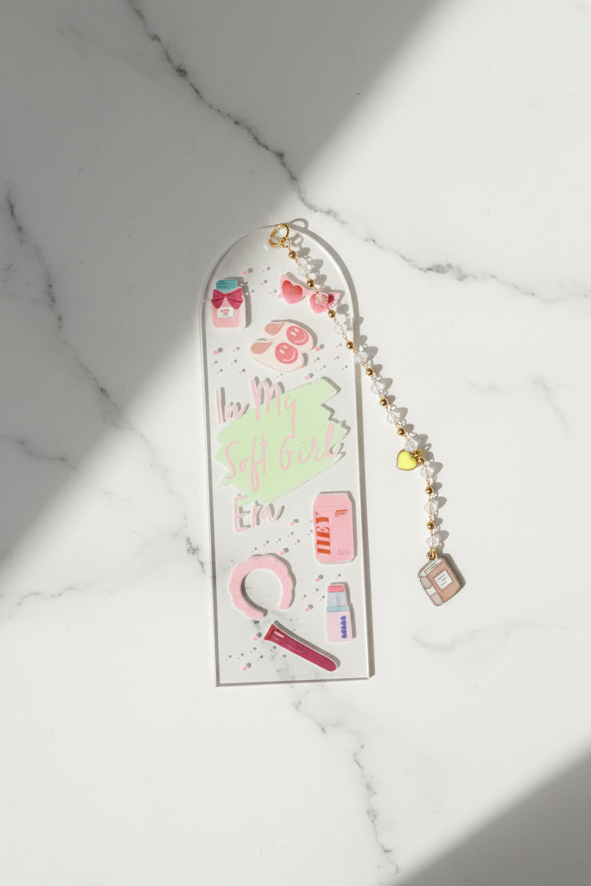 A long, narrow acrylic wall plaque or decorative panel with rounded top corners, featuring pastel illustrations and the phrase 'love is all that matters' in pink script. A beaded gold-and-white chain with small charms is draped beside it on a white marble surface with strong diagonal sunlight and shadows.