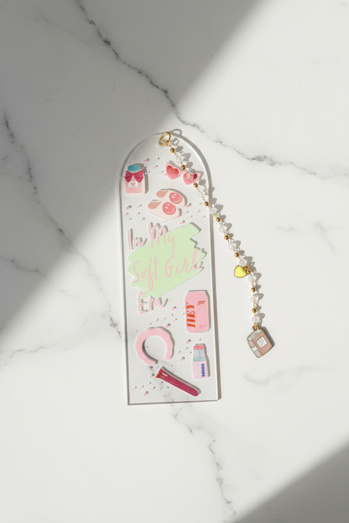 A long, narrow acrylic wall plaque or decorative panel with rounded top corners, featuring pastel illustrations and the phrase 'love is all that matters' in pink script. A beaded gold-and-white chain with small charms is draped beside it on a white marble surface with strong diagonal sunlight and shadows.