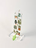 A clear acrylic hanging plaque featuring a botanical illustration of potted succulents and a green watering can, displayed on a clear acrylic stand with a small green leaf charm in front, set against a plain light gray background.