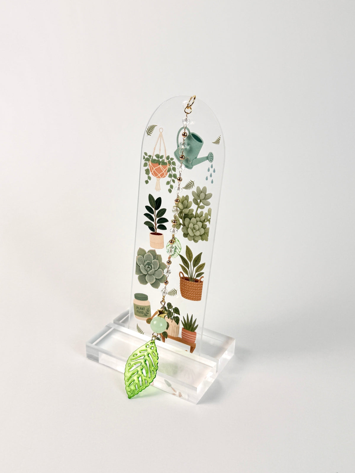 A clear acrylic hanging plaque featuring a botanical illustration of potted succulents and a green watering can, displayed on a clear acrylic stand with a small green leaf charm in front, set against a plain light gray background.