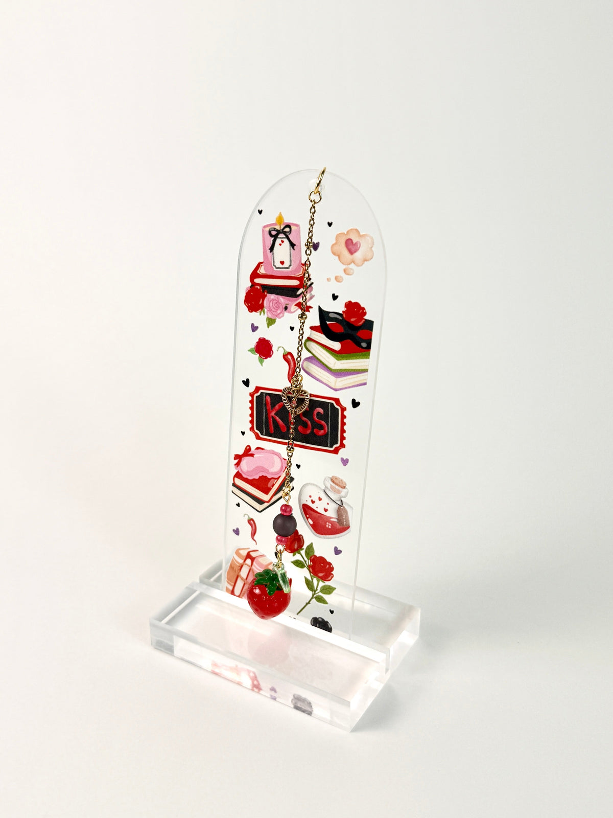 A clear arched acrylic bookmark with red strawberry charm that says KISS in the middle with books, masks, candles and roses sitting on a acrylic stand on a white background. 