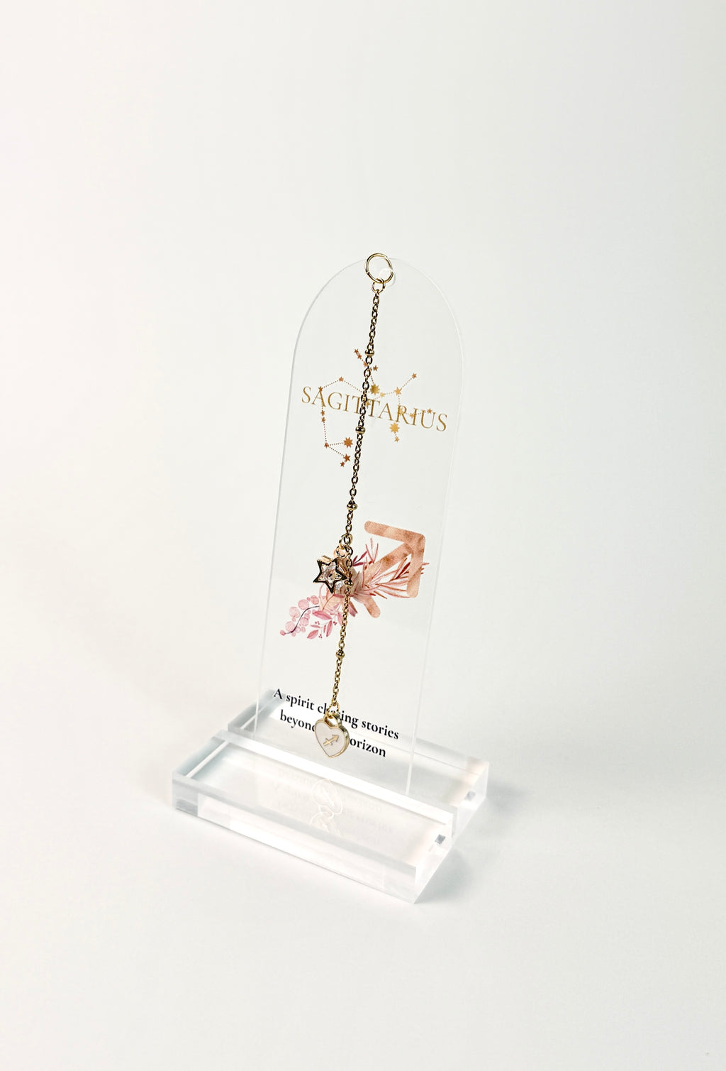 Clear arched acrylic bookmark with Sagittarius zodiac lettering, constellation artwork, an archer symbol with pink botanical details, the quote “A spirit chasing stories beyond the horizon,” and dangling star and Sagittarius heart charms. standing on white background