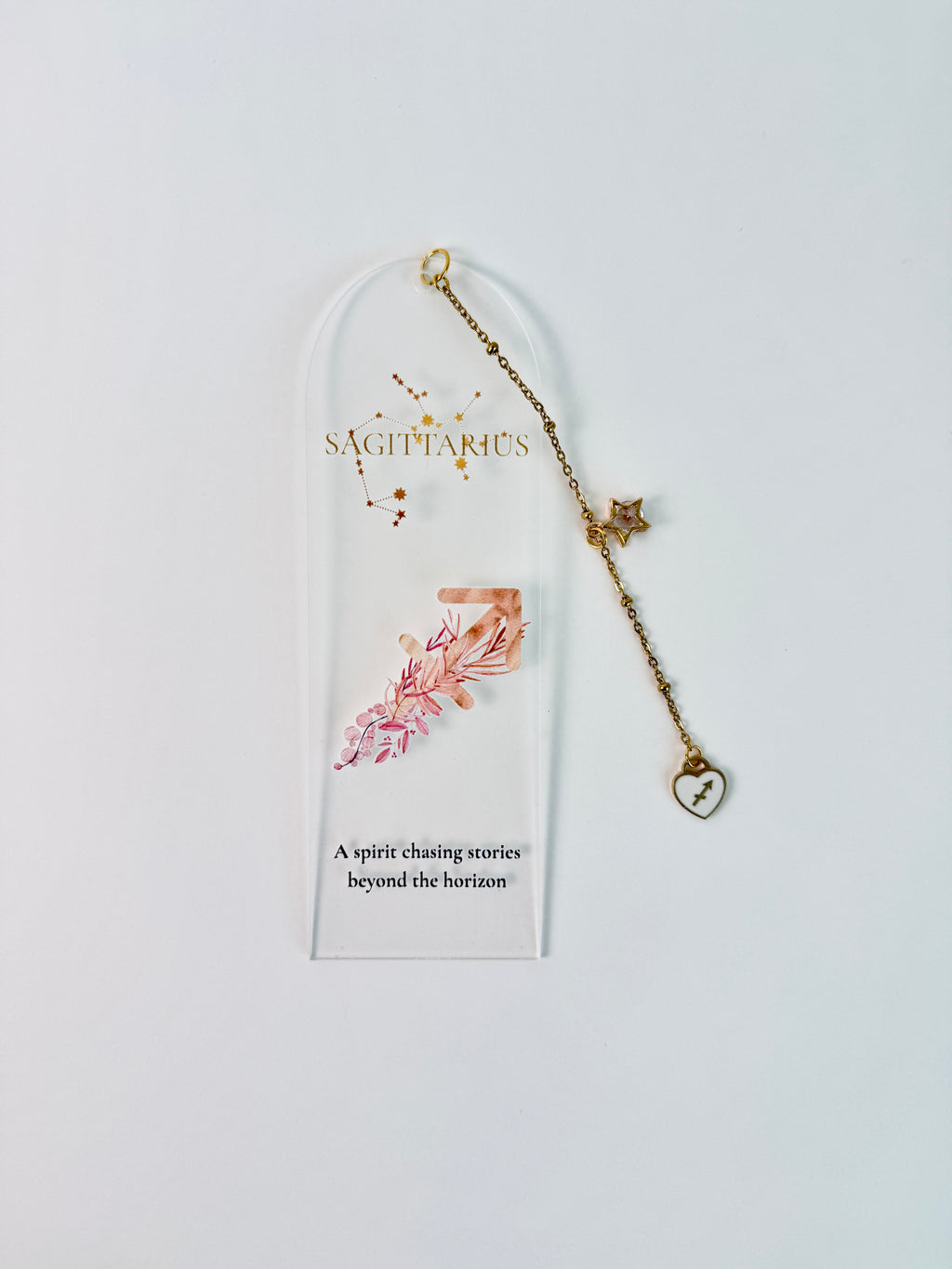 Clear arched acrylic bookmark with Sagittarius zodiac lettering, constellation artwork, an archer symbol with pink botanical details, the quote “A spirit chasing stories beyond the horizon,” and dangling star and Sagittarius heart charms. laying flat on white background