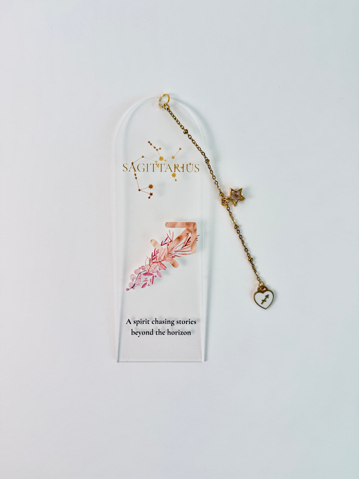 Clear arched acrylic bookmark with Sagittarius zodiac lettering, constellation artwork, an archer symbol with pink botanical details, the quote “A spirit chasing stories beyond the horizon,” and dangling star and Sagittarius heart charms. laying flat on white background