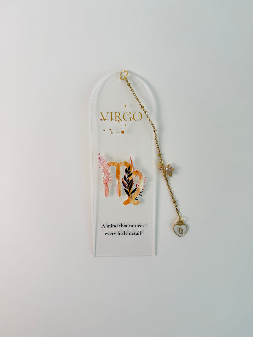 Clear arched acrylic bookmark with Virgo zodiac lettering, constellation artwork, botanical details, the quote “A mind that notices every little detail,” and dangling star and Virgo heart charms. it is laying on white background